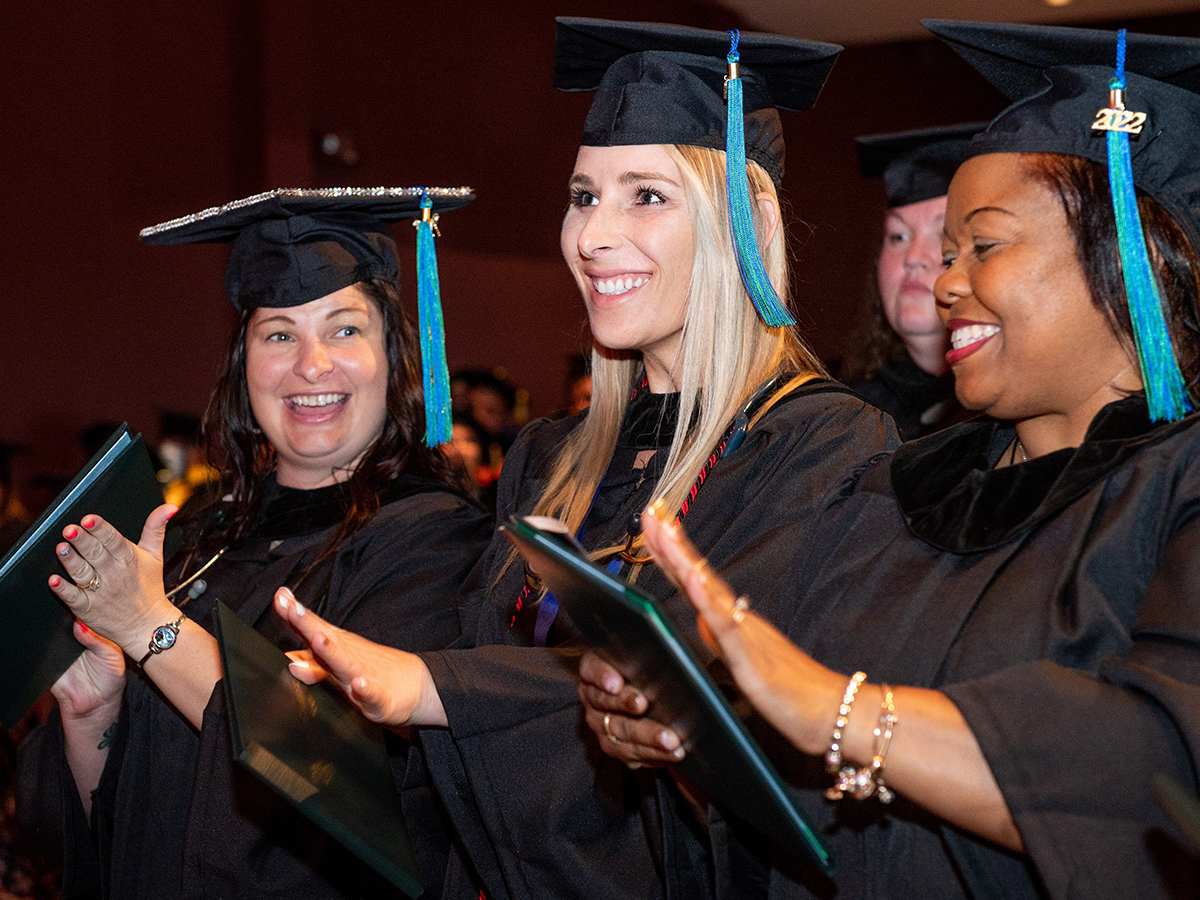 photo of three female graduates in commencement regalia stand side by side and smile and clap their hands