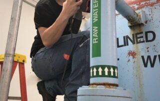 An alumnus crouches on a large pipe next to a pump and smiles for the camera, while holding a large tool in his hand.