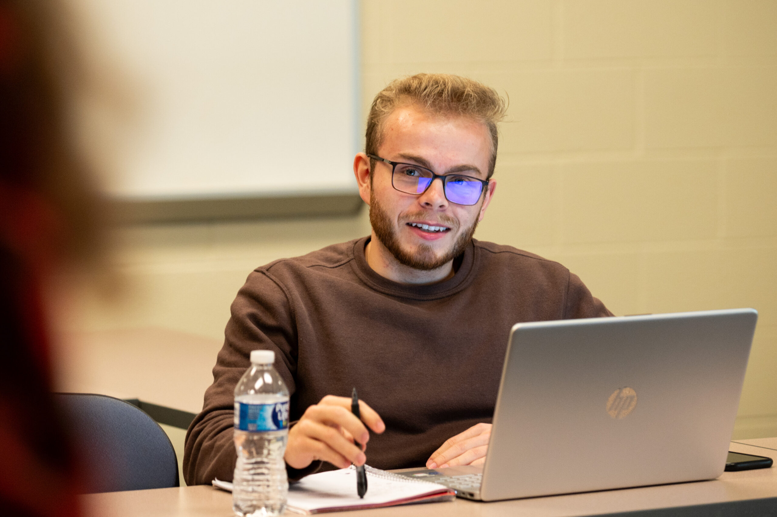 A student with glasses sits in class behind his laptop