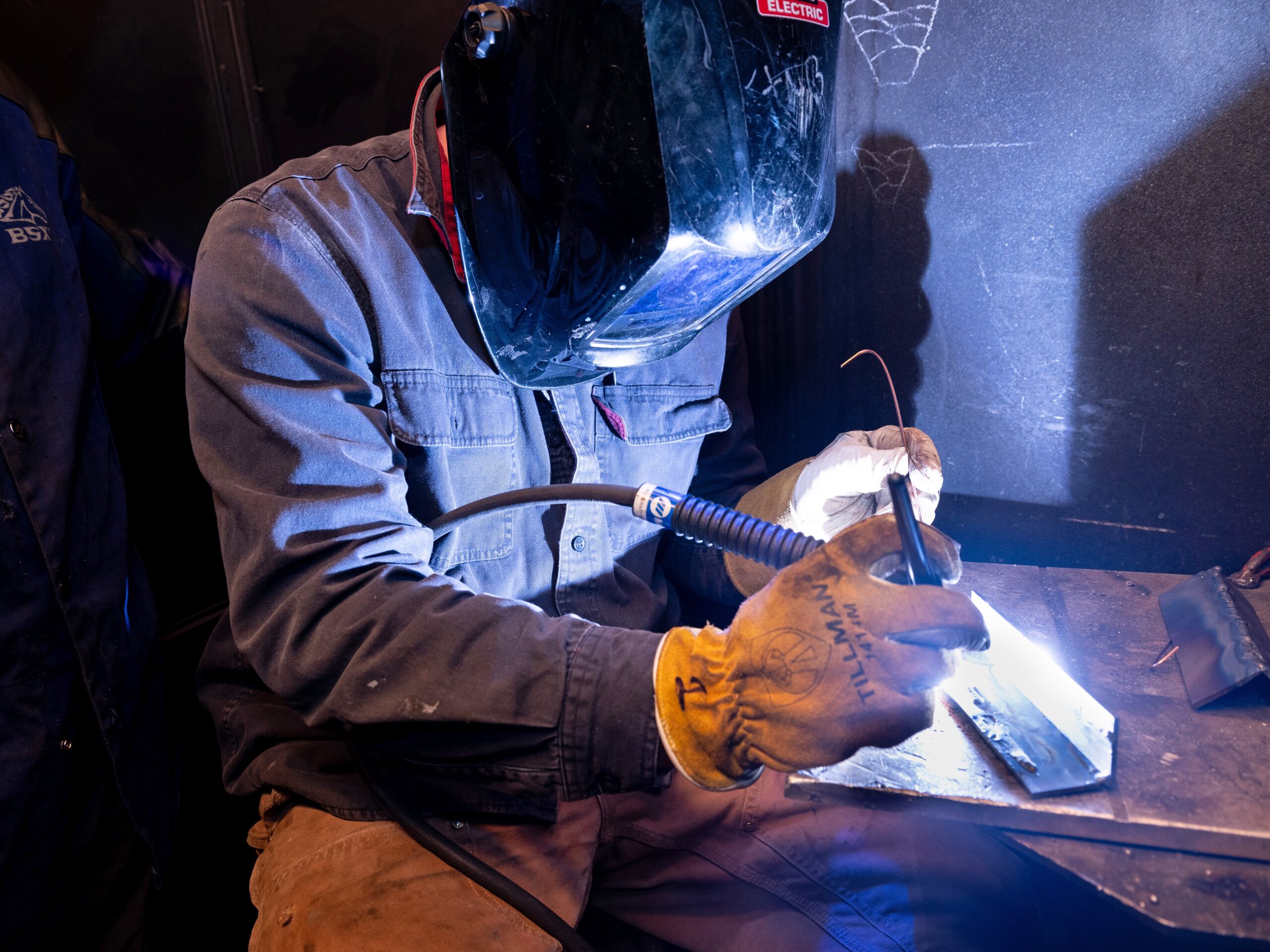 A welding student with shield and gloves works in the shop on a project.