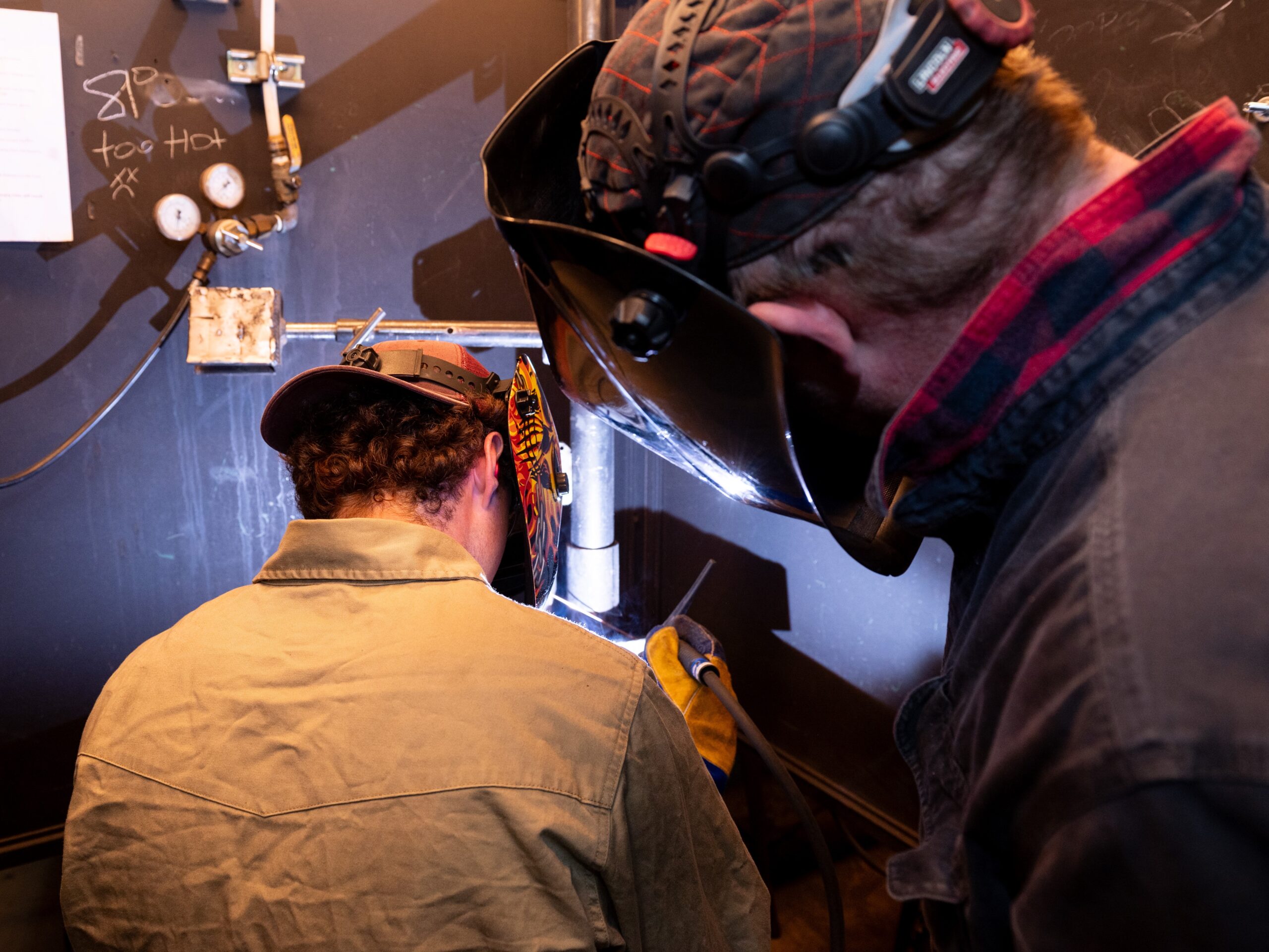 A welding instructor wearing protective gear supervises a student welder.