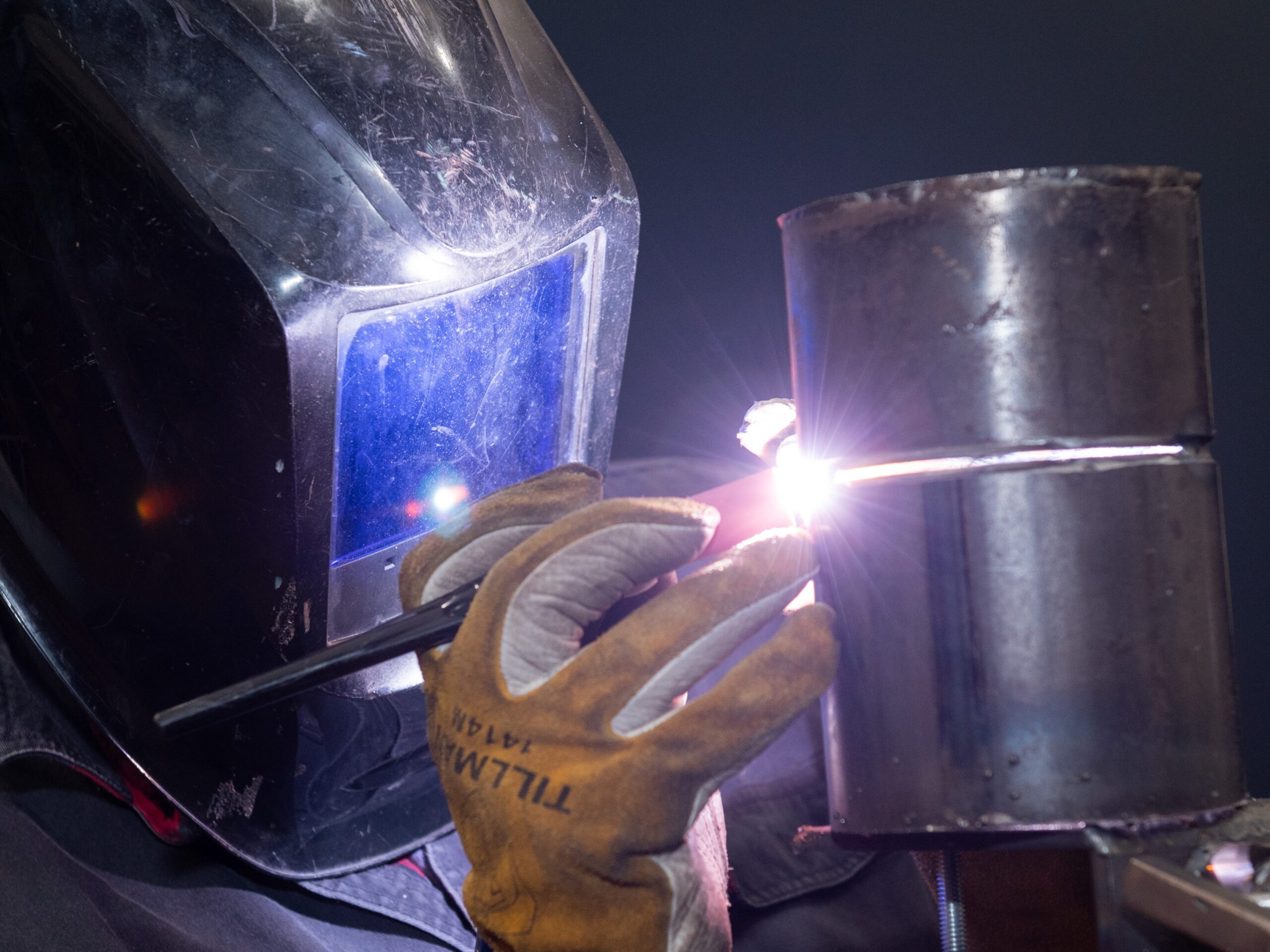 A welder wearing protective gear is working on a metal cylinder.