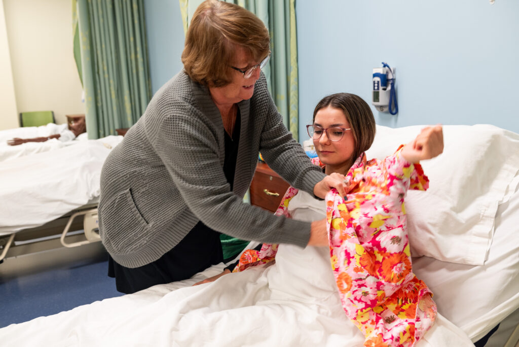 A CNA instructor demonstrates helping a client put on her shirt