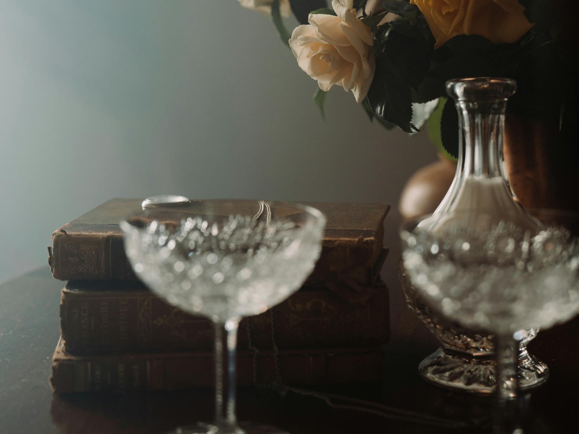 a stack of old books, cut-glass cups and carafe, and faded peach roses in a vase